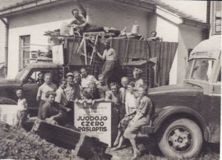 Panevėžio dramos teatro aktoriai ir kiti darbuotojai prie sunkvežimių, pakrautų spektaklio dekoracijomis. Laimutė Liesytė stovi virš afišos, 2-oje eilėje, viduryje. Alytus, 1953 m. PAVB FKV-292-2-1