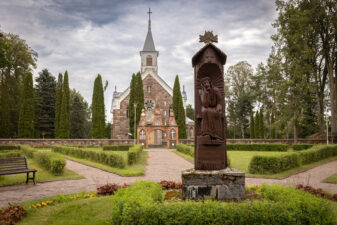 Andrioniškio Šv. apaštalų Petro ir Povilo bažnyčia. Priekyje – 1989 m. pastatytas ąžuolinis Rūpintojėlis (autorius – tautodailininkas Povilas Bugailiškis). Fotogr. G. Lukoševičius. Andrioniškis (Anykščių r.). 2025 m. PAVB skaitmeninis archyvas