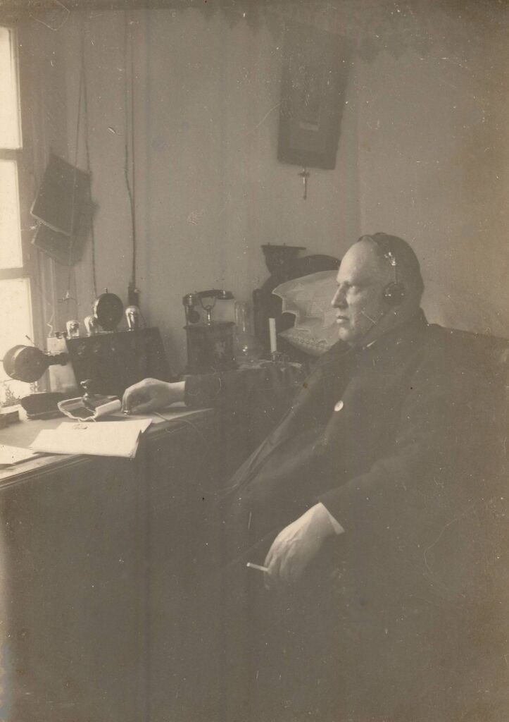 Raguvos Švč. Mergelės Marijos Ėmimo į dangų parapijos klebonas Jonas Maciejauskas savo darbo kambaryje klausosi radijo. Raguva (Panevėžio r.). Apie 1930 m. PAVB, Pavienių rankraščių fondas F8-660-3